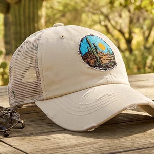 May include: A cream-colored baseball cap with a mesh back and a distressed brim. The front features a circular embroidered patch depicting a desert landscape with a saguaro cactus, brown mountains, and a bright yellow sun against a blue sky.