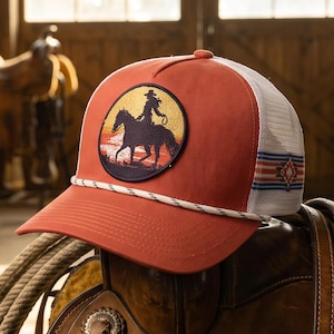 May include: A rust-orange trucker hat featuring a circular patch with a silhouette of a cowboy riding a horse against a sunset background. The hat has a white mesh back, a white rope detail across the brim, and a red, white, and blue striped pattern.