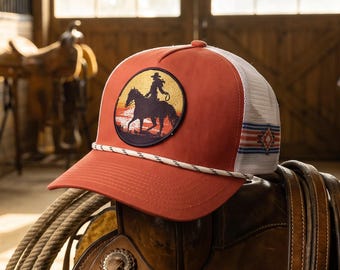 Könnte beinhalten: Eine rostorange Trucker-Cap mit einem runden Aufnäher, der die Silhouette eines Cowboys auf einem Pferd vor einem Sonnenuntergang zeigt. Die Kappe hat ein weißes Netzgewebe an der Rückseite, eine weiße Kordel am Schirm und ein rot-weiß-blaues Streifenmuster.