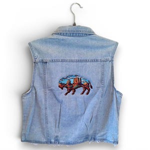 May include: A light blue denim vest featuring a central embroidered patch on the back. The patch depicts a bison silhouette filled with a colorful landscape scene of red rock formations, a blue sky, and green ground, all set against the denim fabric.