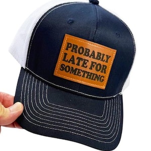 Op de afbeelding: Marineblauwe truckerpet met een witte mesh achterkant. De pet heeft een bruin leren label met de tekst "PROBABLY LATE FOR SOMETHING". De klep is marineblauw met witte stiksels.