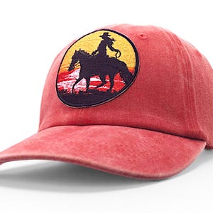 May include: Red baseball cap with a curved brim. The cap features a circular patch with a silhouette of a person riding a horse against a sunset backdrop. The design is in black, yellow, and red.