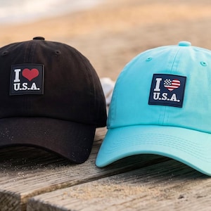 May include: A black baseball cap and a light turquoise baseball cap, each featuring a square patch on the front. The black cap displays a red heart with the text I U.S.A., while the turquoise cap shows a heart with an American flag pattern and the text I U.S.A.
