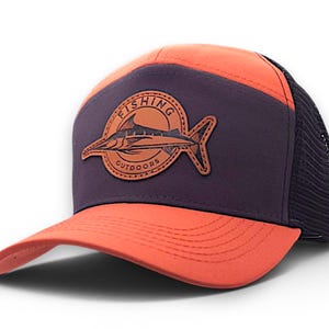 May include: A baseball cap featuring a bright orange brim and top panel, a dark purple front panel, and a black mesh back. A circular tan leather patch on the front displays a graphic of a fish with the text FISHING OUTDOORS in black lettering.
