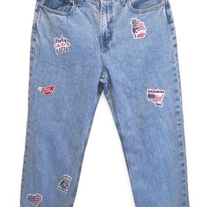 May include: Light blue denim jeans featuring a high-waisted fit and rolled cuffs. The jeans are decorated with various iron-on patches, including text that reads Party Like It's 1776, Independence Day, and Ben Drankin, alongside American flag motifs.