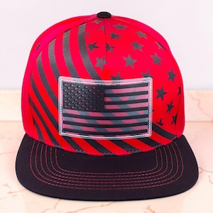 Könnte beinhalten: Eine rot-schwarze Baseballkappe mit einem grafischen Muster aus schwarzen Streifen und Sternen auf der Krone. Ein rechteckiger Aufnäher mit einer schwarzen amerikanischen Flagge ist mittig auf der Vorderseite angebracht. Die Kappe hat einen flachen schwarzen Schirm mit roten Nähten.