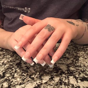 Op de afbeelding: Een set lange, vierkant gevormde kunstnagels met een klassiek French manicure-ontwerp met een transparante, natuurlijk getinte basis en helderwitte uiteinden. De nagels zijn in een overlappende positie gerangschikt.