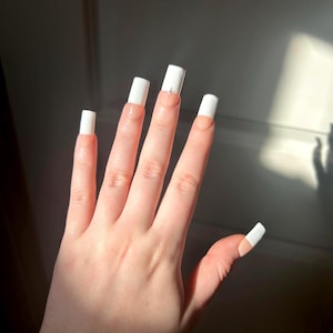 May include: A set of long, square-shaped artificial nails featuring a classic French manicure design with opaque white tips and a natural-toned base. One nail is accented with a small, silver-toned star-shaped charm placed at the center of the tip.
