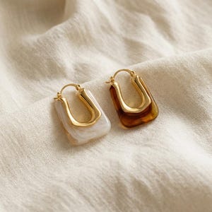 May include: A pair of hoop earrings featuring a U-shaped design. One earring has a white marbled resin base with a gold-toned metal insert, while the other has a translucent brown marbled resin base with a matching gold-toned metal insert.