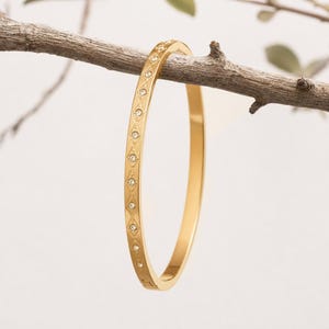 May include: A gold-colored bangle bracelet with a series of small, clear gemstones set in a repeating pattern. The bracelet is displayed on a brown branch against a soft, neutral background. The bangle is a simple, elegant piece of jewelry.