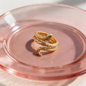 May include: A gold-colored snake ring with a textured, scale-like design. The snake's head features a small green gemstone eye. The ring is coiled on a pink, glass plate, creating a striking contrast.