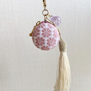 May include: A pink and white embroidered round charm with a gold chain and tassel. The charm has a floral pattern and a small purple bead. The tassel is cream-colored and hangs from the bottom.