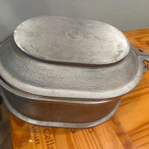 May include: An oval-shaped silver-toned metal roasting pan with a textured exterior finish and a smooth, flat lid. The pan features integrated side handles with a distinct notched design on each end, providing a secure grip for lifting.