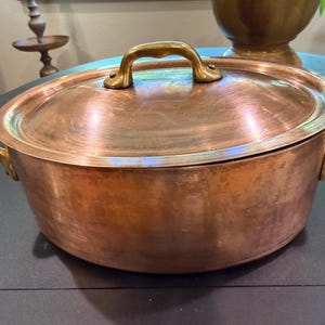 Pode incluir: Uma panela de cozinha de cobre com tampa. A panela tem duas alças laterais e uma alça superior, todas em dourado. A panela é redonda e assenta sobre uma superfície escura. É feita de cobre e tem um acabamento brilhante.