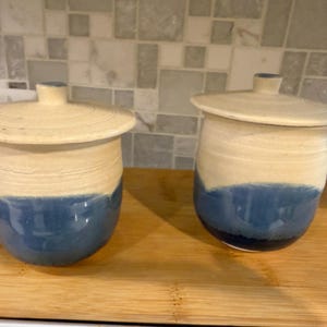 May include: Two ceramic jars with lids. The jars are cylindrical with a cream-colored top and a blue bottom. The lids are also cream-colored. The jars are sitting on a wooden cutting board.