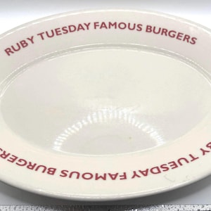May include: Off-white oval serving platter with the text "RUBY TUESDAY FAMOUS BURGERS" in red lettering around the rim. The platter has a glossy finish and is likely made of ceramic or porcelain.
