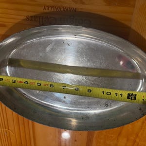 May include: An oval-shaped silver-toned metal serving platter with two side handles. A yellow measuring tape is stretched across the center of the platter, indicating a length of approximately 14 inches from edge to edge.