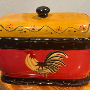 May include: A rectangular ceramic butter dish with a red base featuring a painted rooster, a black scalloped rim with gold dots, and a yellow lid decorated with orange flowers and a black knob handle on top.