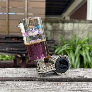 May include: A cylindrical gear shift knob featuring a clear resin top containing a small purple cartoon character figure with orange hair, set above a layer of gold flakes and a solid purple base, attached to a polished chrome metal mounting bracket.