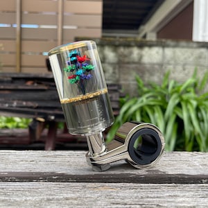 May include: A cylindrical gear shift knob made of clear resin containing a bouquet of small red, blue, and green dried flowers above a layer of gold flakes, attached to a polished chrome metal mounting bracket with a circular clamp mechanism.