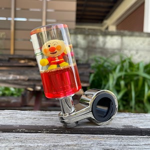 May include: A cylindrical gear shift knob featuring a clear acrylic top section containing a small Anpanman character figure and gold glitter, with a vibrant red base. The knob is attached to a polished chrome metal mounting bracket with a black ring.
