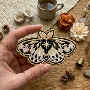 Puede incluir: Parche de polilla bordada con un contorno negro y dorado, con una estrella y detalles florales. Las alas de la polilla tienen una combinación de colores crema y rosa. El parche está sostenido por una mano, con otros suministros de artesanía en el fondo.