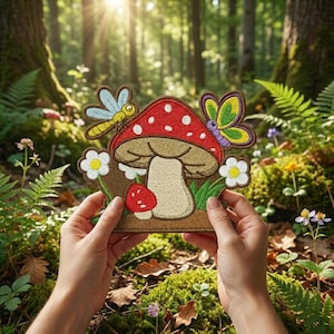 May include: An embroidered patch featuring a large red mushroom with white spots, a smaller red mushroom, a yellow dragonfly, a purple and green butterfly, and two white flowers with yellow centers, all set against a brown background.