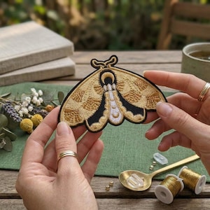 Puede incluir: Parche de polilla bordada en oro y negro. El parche presenta intrincadas puntadas, mostrando las alas y el cuerpo de la polilla. Se sostiene sobre un fondo verde, con una cuchara, carretes de hilo y otros objetos.