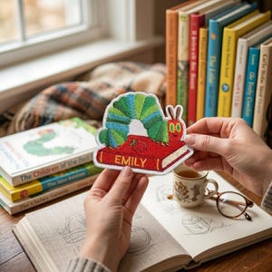 Peut inclure: Un écusson brodé coloré représentant une chenille verte et bleue sur un livre rouge avec le nom "EMILY". L'écusson est tenu près d'une pile de livres pour enfants et d'une tasse de thé.