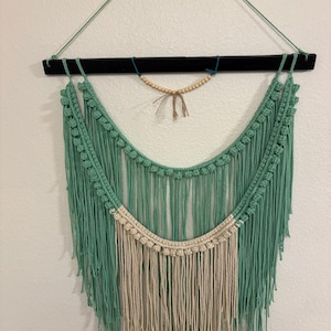 May include: A macrame wall hanging featuring a teal and cream color scheme. The top is a black wooden bar with a teal string for hanging. The design includes a beaded accent and long, fringed tassels.