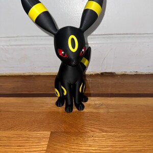 May include: A black figurine of a Pokemon character with yellow accents on the ears, legs, and tail. The figurine has red eyes and a yellow ring on its forehead. The character is sitting on a wooden surface.