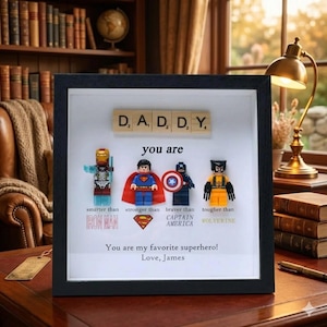 May include: A framed artwork featuring the word "DADDY" spelled out with Scrabble tiles. Below, Lego superhero figures of Iron Man, Superman, Captain America, and Wolverine are displayed with text. The bottom reads "You are my favorite superhero! Love, James."