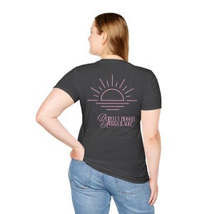 Op de afbeelding: Een donkergrijs t-shirt met korte mouwen en een lichtroze afbeelding van een opkomende zon op de achterkant. Onder de zon staat de tekst "DOPO LA PIOGGIA ARRIVA IL SOLE" in een gestileerd roze lettertype. Het t-shirt wordt gedragen met een blauwe spijkerbroek.