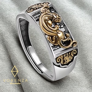 Puede incluir: Un anillo plateado con detalles dorados, que presenta un diseño ornamentado. La parte superior rectangular del anillo presenta un motivo dorado en forma de dragón y caracteres chinos. El anillo es de Vorenza Silver Atelier.