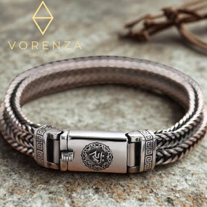 Könnte beinhalten: Ein silbernes Armband mit geflochtenem Design und einem rechteckigen Verschluss. Der Verschluss hat ein eingraviertes kreisförmiges Emblem und geometrische Muster. Das Armband wird auf einer strukturierten Oberfläche präsentiert, mit dem Markennamen "Vorenza".