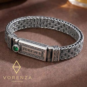 Könnte beinhalten: Ein silbernes Armband mit geflochtenem Kettendesign und einem rechteckigen Verschluss, in den das Wort FOREVER eingraviert ist. Der Verschluss ist mit filigranen Schnörkeln und einem einzelnen runden grünen Edelstein an der Seite verziert.