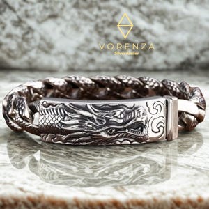 Könnte beinhalten: Silberarmband mit detailliertem Drachenkopf-Verschluss. Das Armband hat ein geflochtenes Band und den Text "VORENZA SilverAtelier". Die Schuppen und Merkmale des Drachen sind aufwendig geschnitzt.