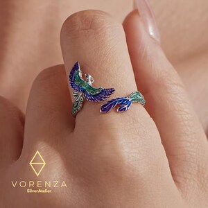 May include: A silver ring with a phoenix design. The ring features a colorful enamel finish in shades of blue, green, and purple. The ring is open, with the phoenix wrapping around the finger. The brand name "VORENZA SilverAtelier" is visible.