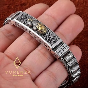 Puede incluir: Pulsera de plata con un diseño detallado. La pulsera presenta una pieza central rectangular con una figura de animal dorada y patrones intrincados. La pulsera está hecha de eslabones de metal trenzado. La marca "Vorenza SilverAtelier" es visible.