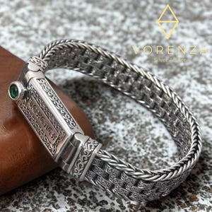 Puede incluir: Una pulsera de plata con un intrincado diseño tejido. La pulsera presenta un cierre rectangular con detalles grabados y una pequeña piedra preciosa verde. La marca "Vorenza Silver Atelier" es visible en el fondo.