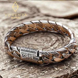 May include: A silver bracelet with an intricate, braided design. The bracelet has a decorative clasp with engraved floral patterns. The brand name "Vorenza SilverAtelier" is visible. The bracelet is displayed on a wooden surface.