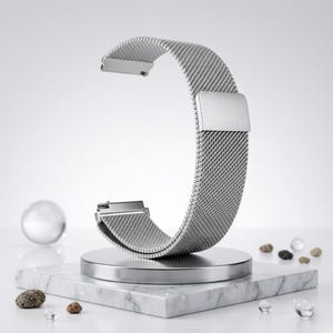 May include: A silver stainless steel watch band with a mesh design. The band is curved and displayed on a small, round, silver stand atop a white marble block. Small rocks and clear spheres are scattered around the base.