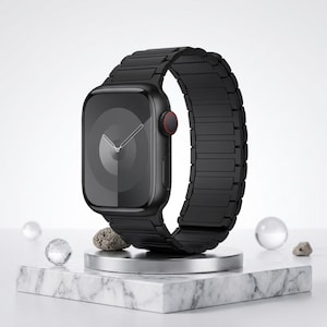 May include: A black smartwatch with a matching black metal band. The watch face displays a minimalist design with white hands. The watch is displayed on a marble block with decorative stones and clear spheres.