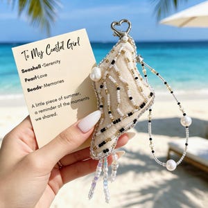 May include: A cream-colored fabric charm shaped like a seashell, embellished with black, gold, and clear beads, featuring a heart-shaped clasp and a beaded lanyard with pearls. It is accompanied by a card reading To My Coastal Girl.