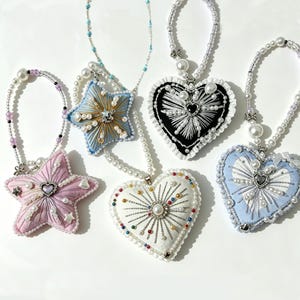 May include: A collection of five hanging ornaments in star and heart shapes, crafted from fabric with embroidered sunburst patterns, central heart charms, and beaded pearl loops. The ornaments feature pastel pink, light blue, white, and black colors.