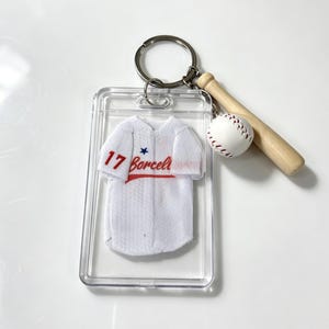 May include: A keychain featuring a clear rectangular plastic case containing a miniature white baseball jersey with the number 17, a blue star, and the red text Borcelli. Attached is a small wooden baseball bat and a white baseball charm.