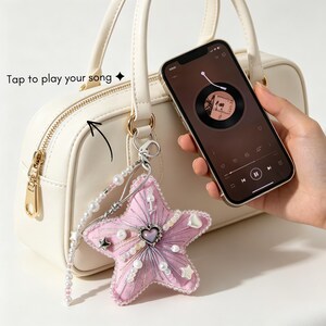 Puede incluir: Un bolso de color crema con un llavero en forma de estrella de tela rosa, costuras plateadas, perlas blancas y un corazón central. El llavero está unido a una anilla plateada. Un smartphone muestra un reproductor de música con el texto Tap to play your song.