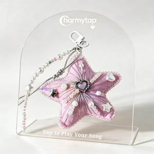 May include: A pink star-shaped plush charm featuring a central heart-shaped charm, silver metallic accents, and white bead embellishments. It hangs from a beaded strap with a silver clasp, displayed on a clear acrylic stand with the text Charmytap and Tap to Play Your Song.