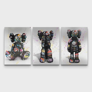 May include: Triptych of canvas prints showcasing black, graffiti-style figures. Each figure is in a different pose and adorned with vibrant text and patterns. The background is white with colourful paint splatters.