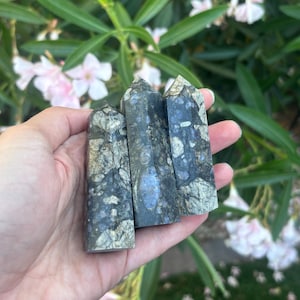 Pode incluir: Três torres de cristal polido com formato hexagonal, apresentando uma cor base cinza-escura e preta com inclusões metálicas de pirita dourada e manchas de labradorita azul iridescente em todas as superfícies de pedra.